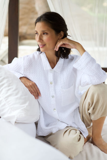 A woman sitting on a bed in a Waffle Linen Pocket Shirt and tan pants.