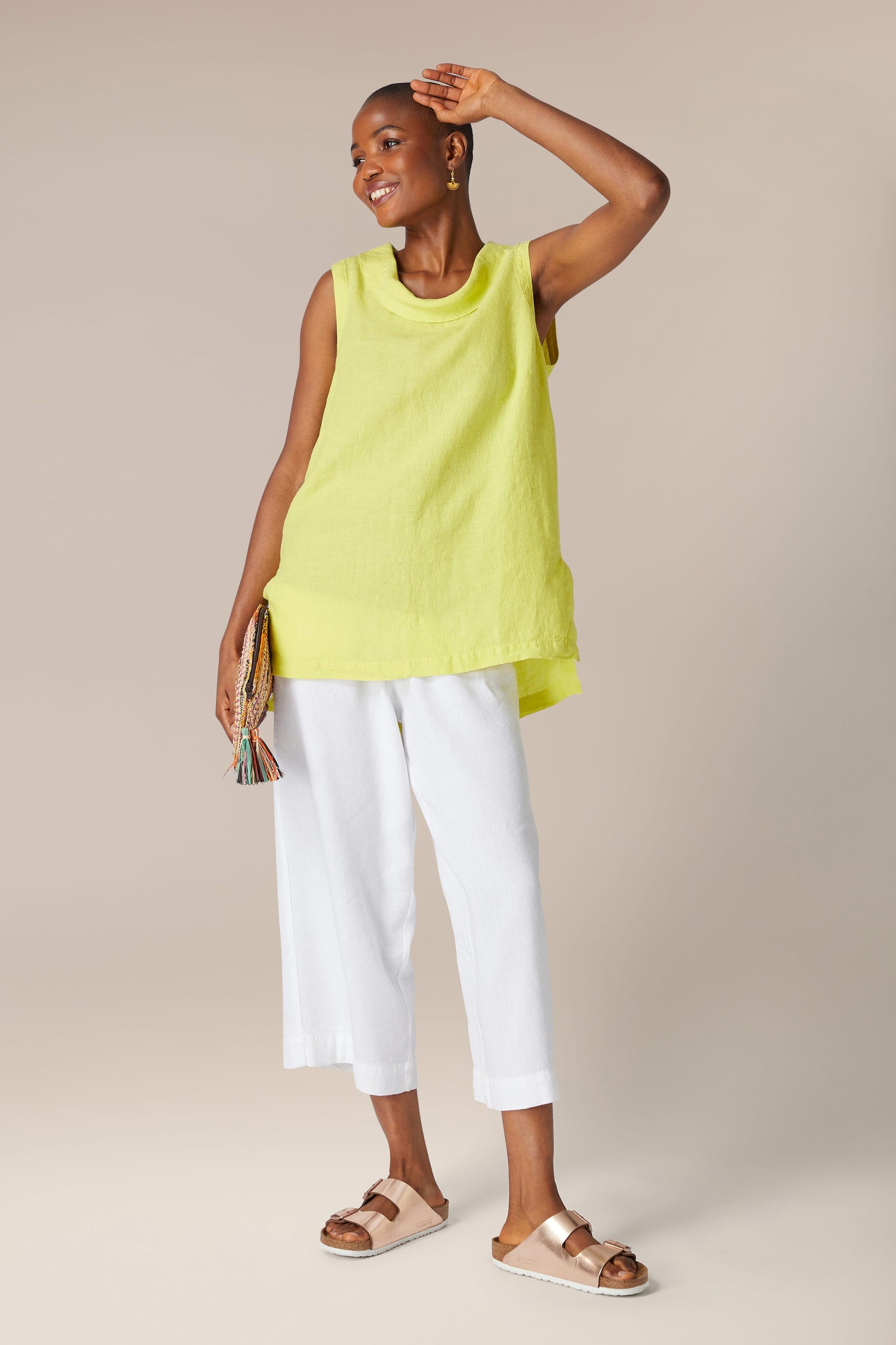 A smiling woman in a Organza Linen Cowl Vest, white capri pants, and beige sandals, holding a multicolored clutch, standing against a neutral background.