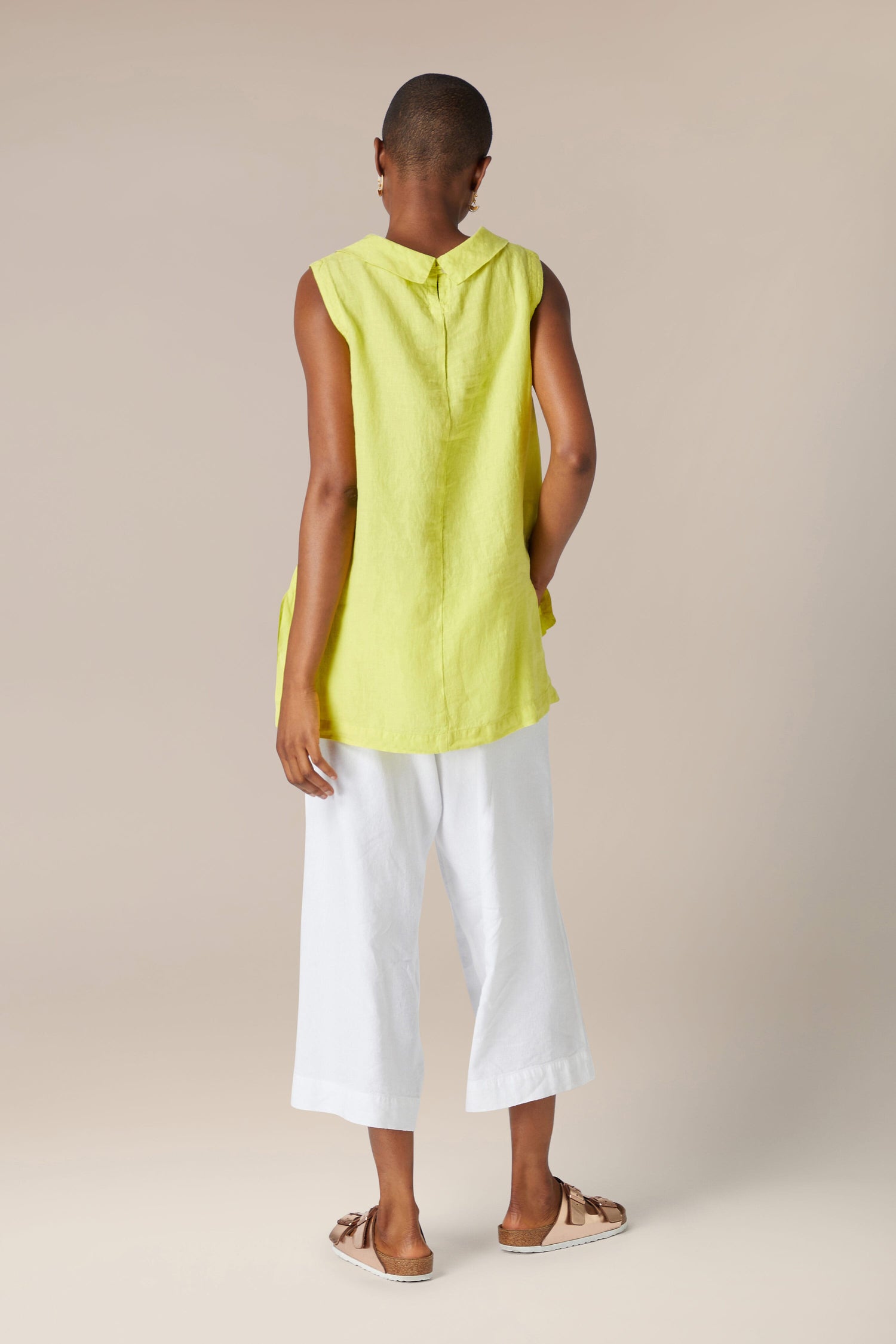 A woman stands facing away from the camera, wearing an Organza Linen Cowl Vest and white cropped trousers, in a neutral-colored room.