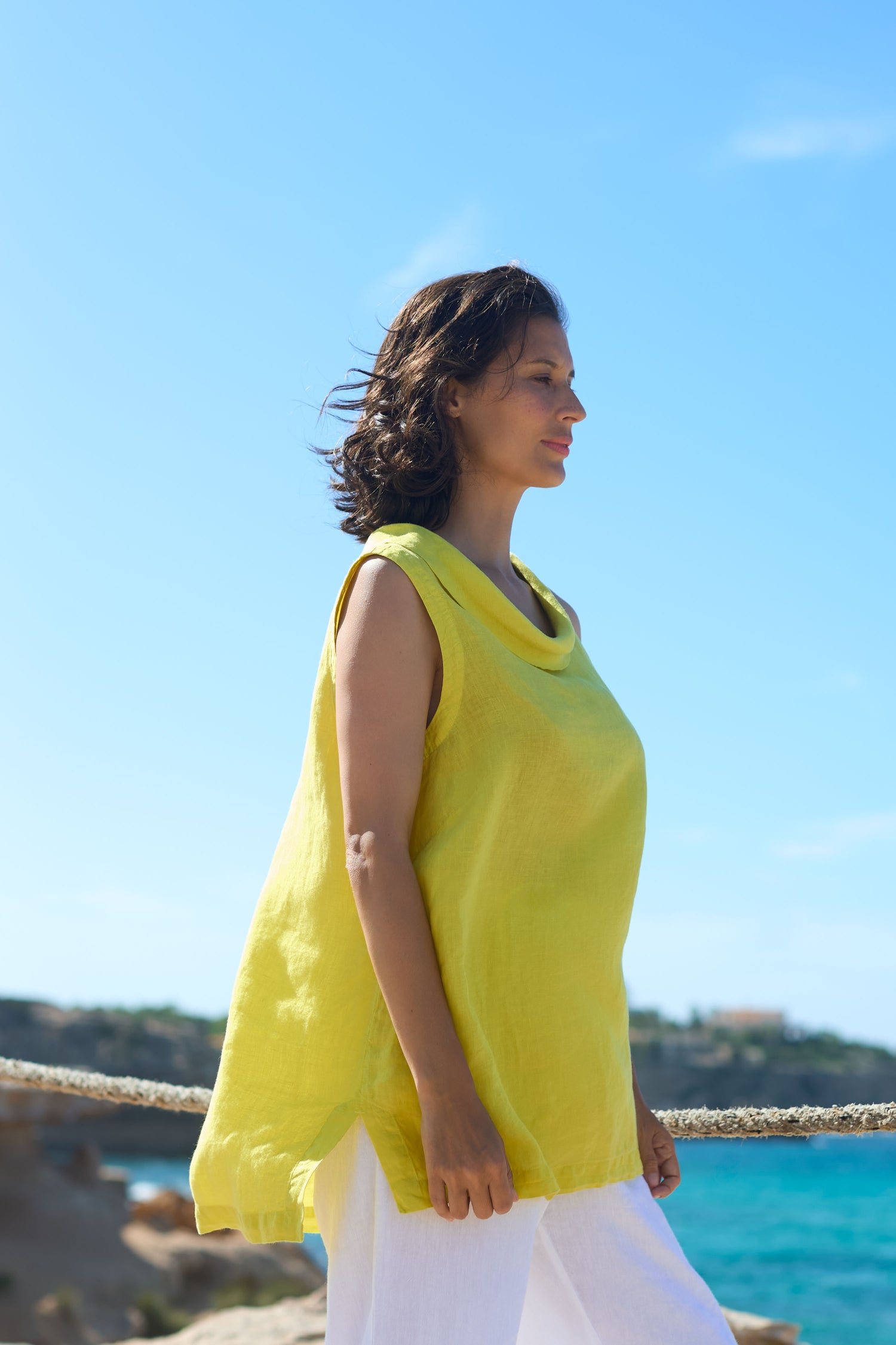 Woman in an Organza Linen Cowl Vest yellow top and white pants standing by the seaside, with a serene expression, looking to her side.