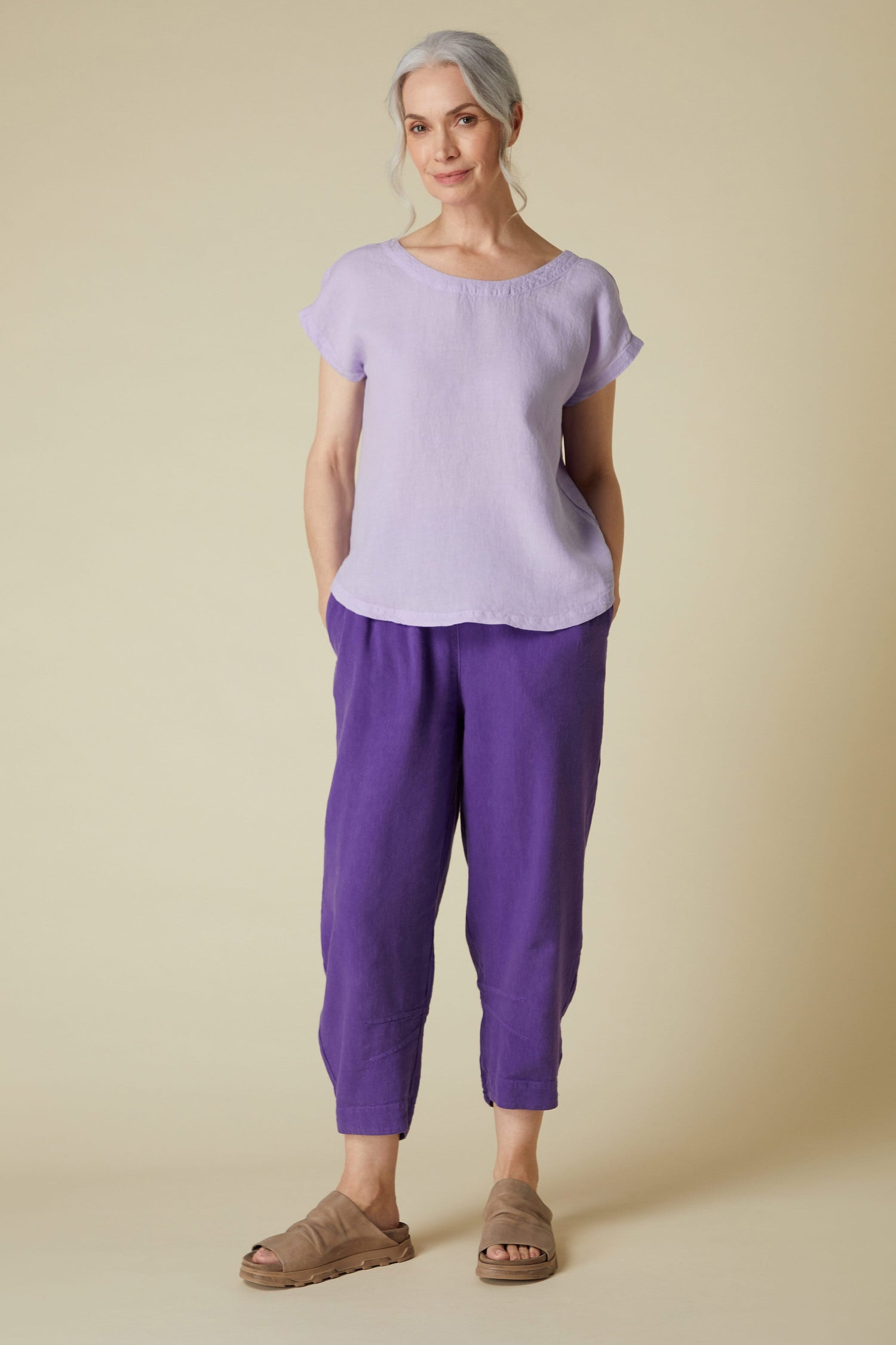 A woman stands posing in a Organza Linen Reversible Top and dark purple trousers with beige sandals.