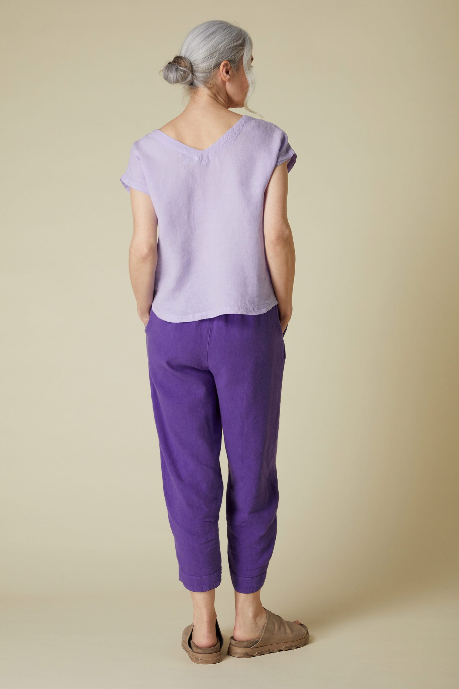 Woman standing with her back to the camera, dressed in a Organza Linen reversible, short-sleeved top and purple pants, with her hair in a bun.