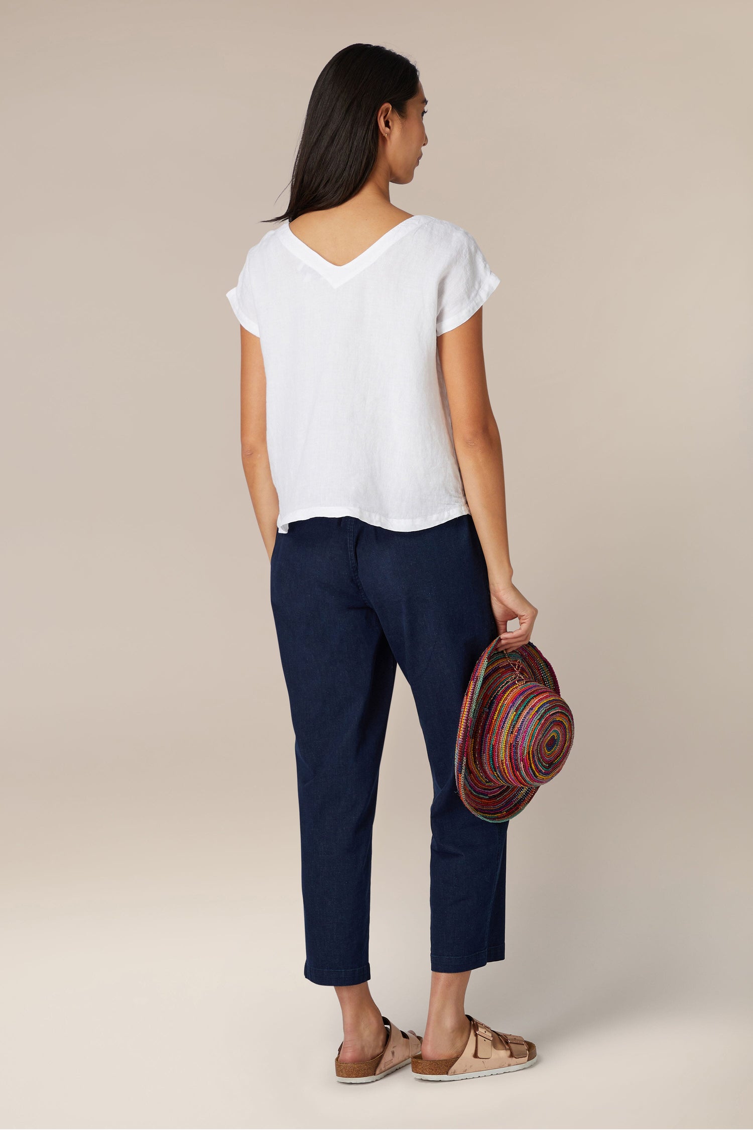 A woman standing with her back to the camera, holding a colorful hat, wearing an Organza Linen Reversible Top, navy crop pants, and patterned sandals.