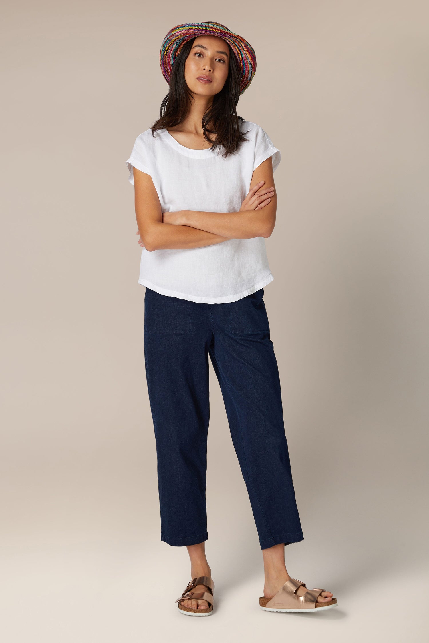 Sentence with Product Name: A woman in a white Bella Organza Linen Reversible Top and navy cropped pants with a colorful hat stands confidently against a neutral background.