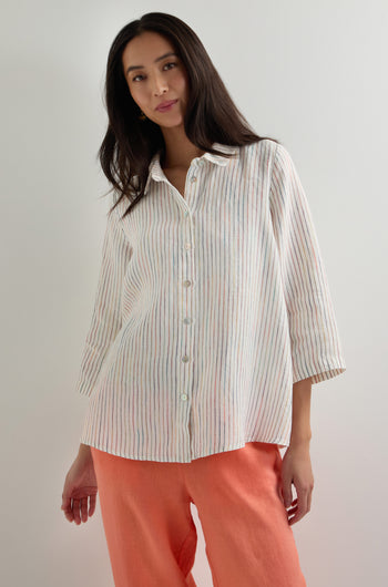 A woman stands against a plain background wearing a White Space Dye Stripe Linen Shirt and coral-colored pants.