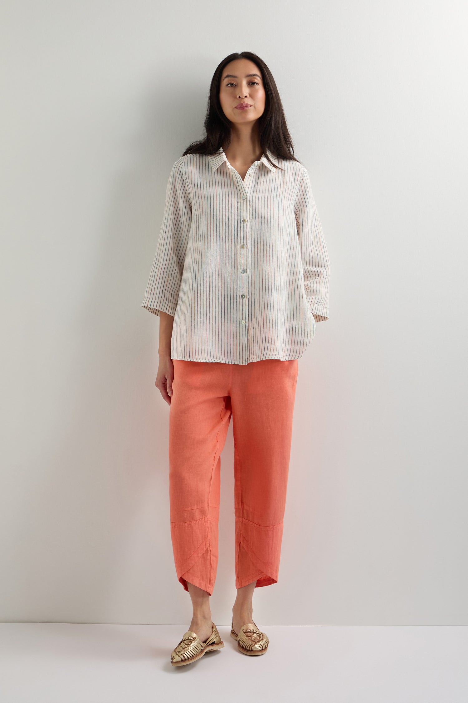 A woman stands against a plain wall wearing a classic fit White Space Dye Stripe Linen Shirt, coral cropped pants, and gold woven slip-on shoes.