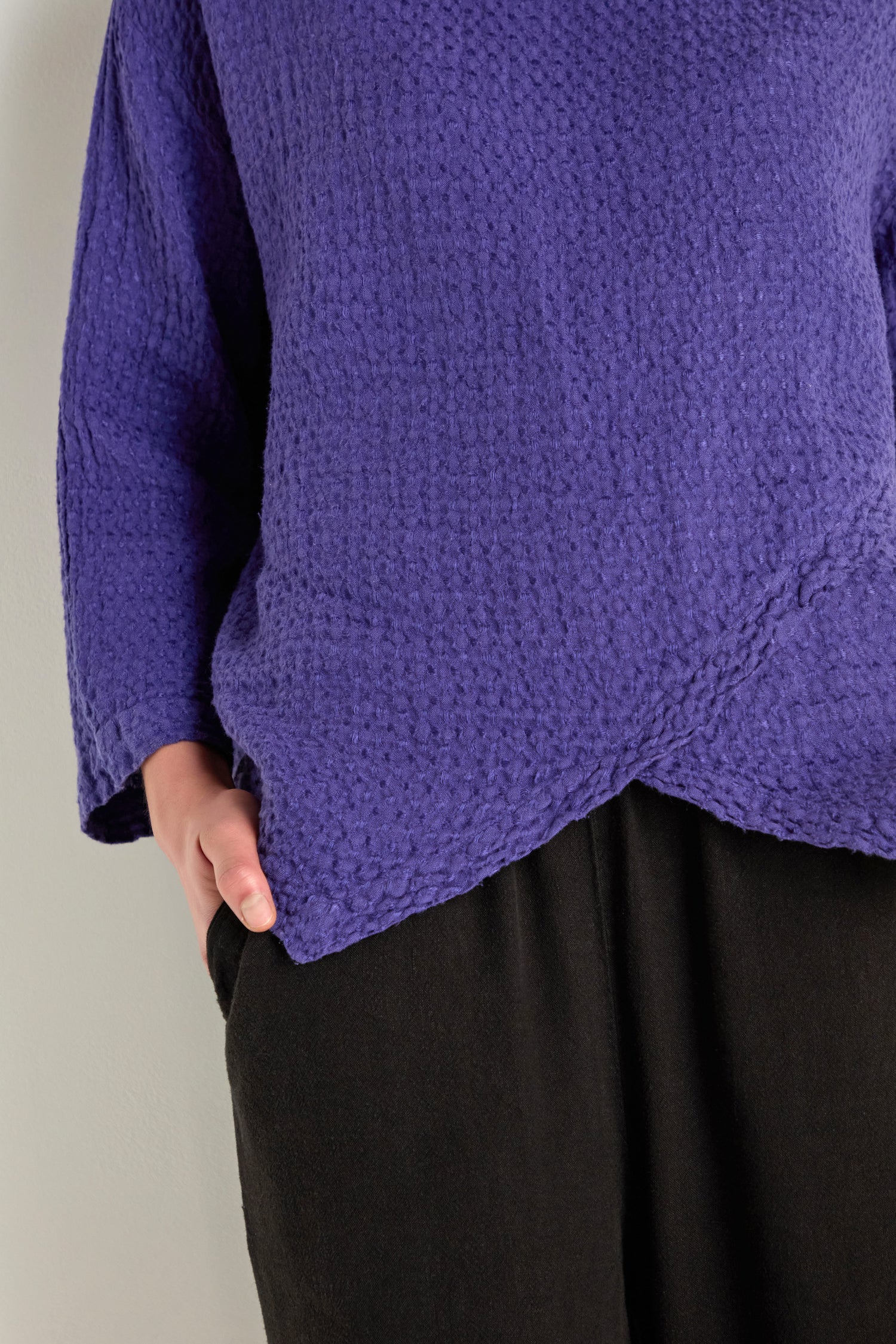 A person wears the Waffle Linen Boxy Cowl Neck Top in textured purple with black pants, one hand in their pocket, standing against a plain light-colored wall.