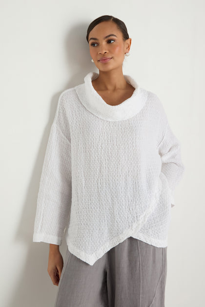 A person stands against a white wall wearing the Waffle Linen Boxy Cowl Neck Top and loose-fitting grey pants.