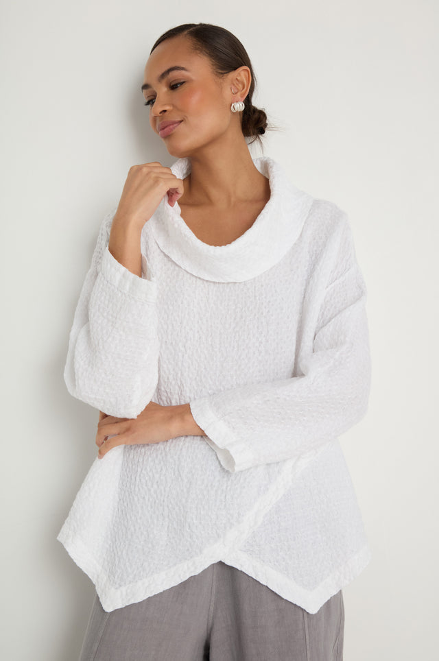 A person stands against a white wall wearing the Waffle Linen Boxy Cowl Neck Top, a textured white sweater with long sleeves, paired with light gray pants.