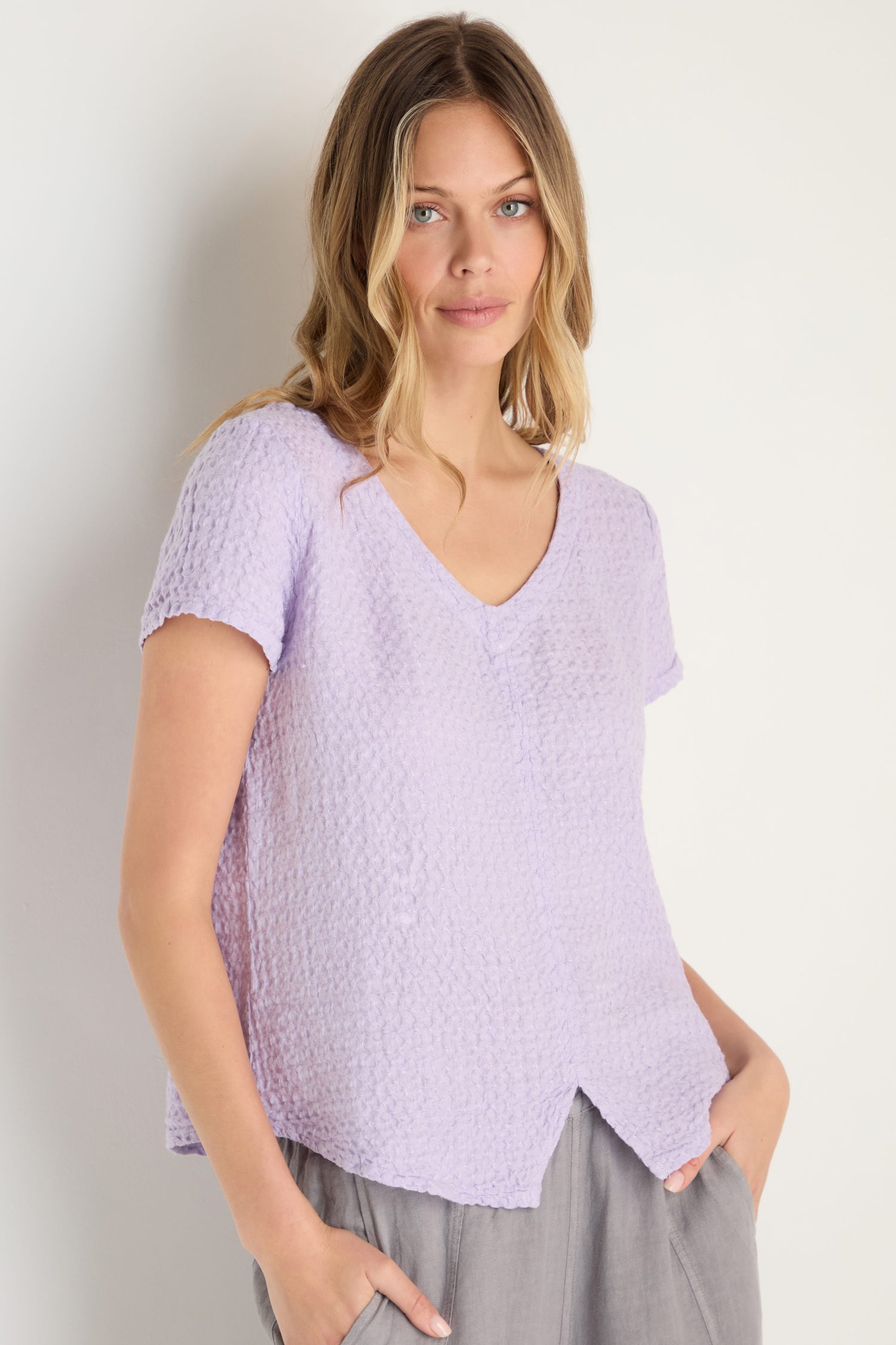 Woman stands against a white wall in the Waffle Linen Lilac V Neck Top and gray pants, one hand in her pocket for a relaxed, breathable look.