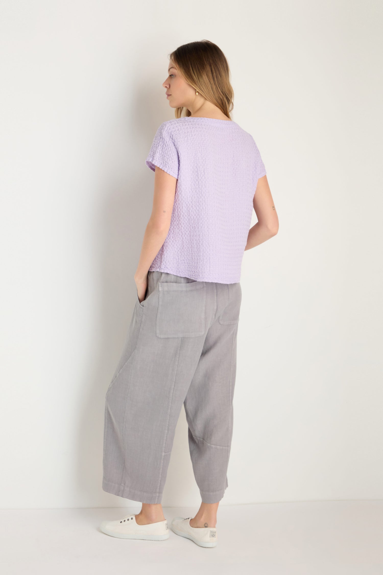 A woman stands facing away, wearing the Waffle Linen Lilac V Neck Top, loose gray pants with pockets, and white sneakers against a plain light background.
