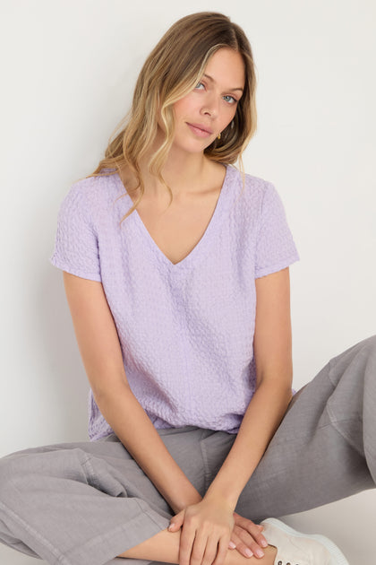 A woman with long blonde hair wears the Waffle Linen Lilac V Neck Top and gray pants while sitting against a white background.
