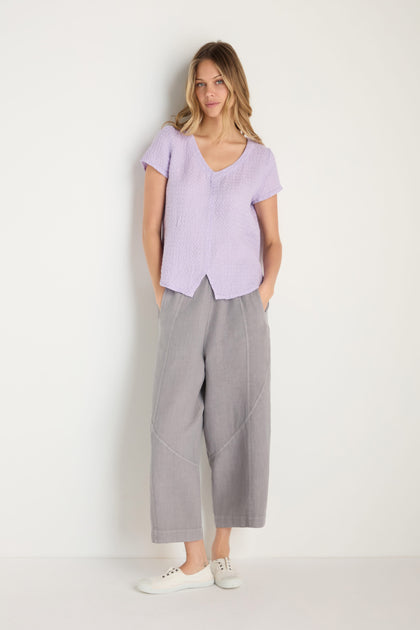 A woman stands against a white wall, hands in her pockets, wearing the Waffle Linen Lilac V Neck Top, loose gray pants, and white sneakers.