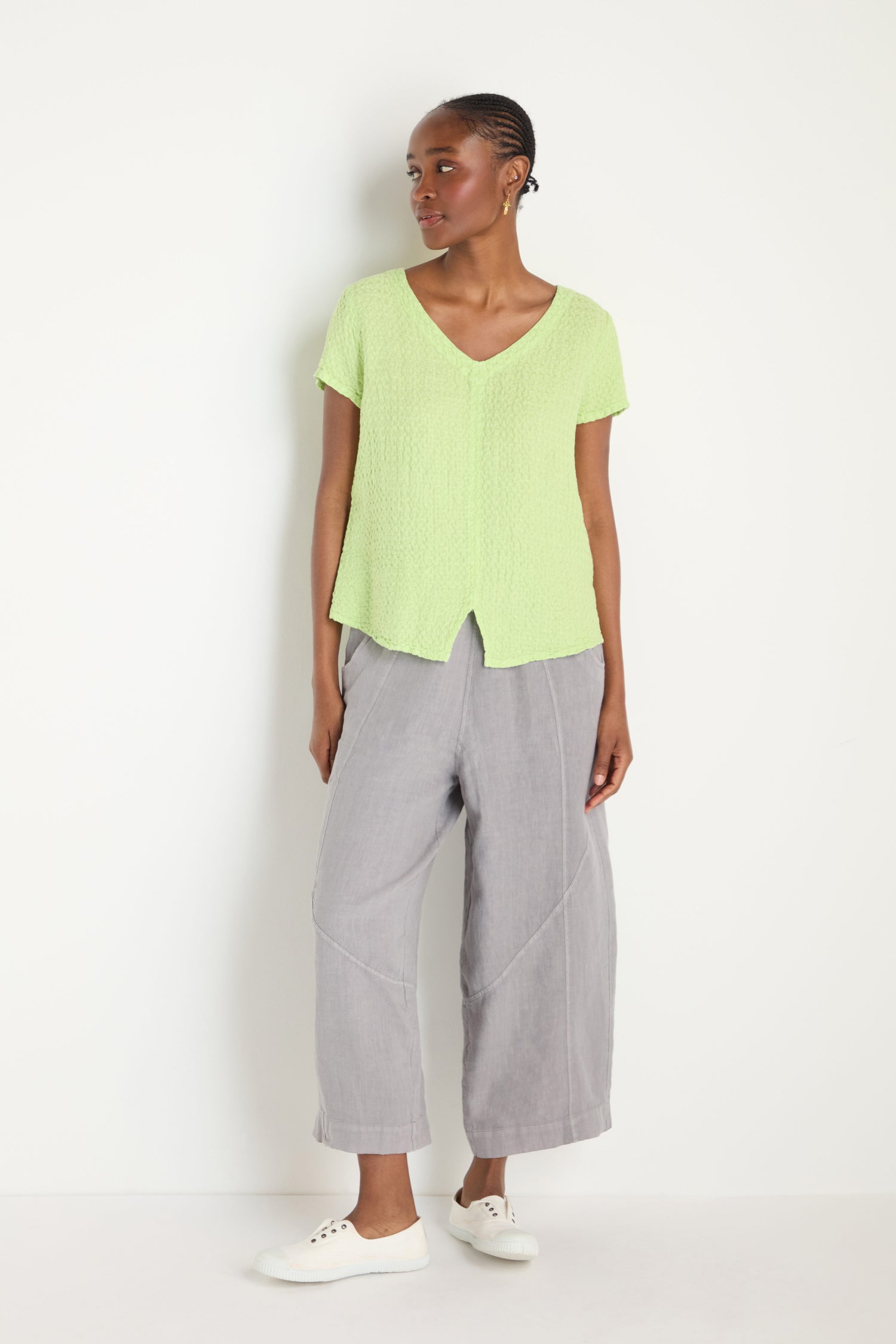 A person stands against a white wall, wearing the Waffle Linen Lime V Neck Top, loose gray pants, and white sneakers.