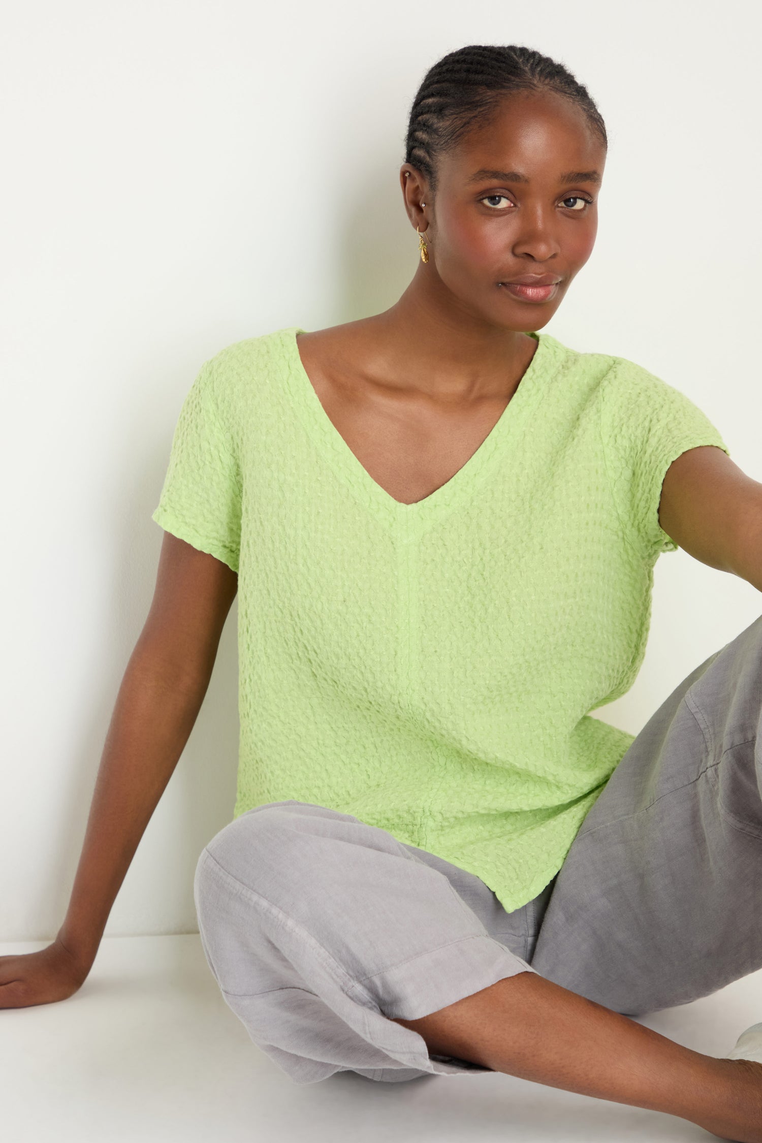 A woman wearing the Waffle Linen Lime V Neck Top and light gray pants sits on the floor against a white background.