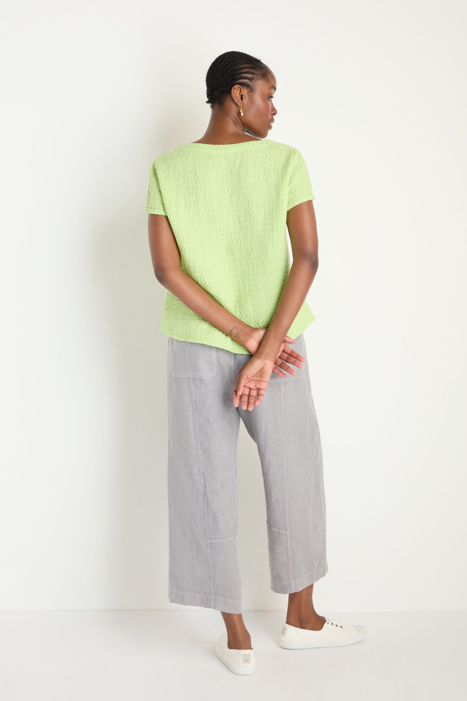 A woman stands against a white wall, facing away, wearing the Waffle Linen Lime V Neck Top, loose gray pants, and white sneakers. Her hands are clasped behind her back.