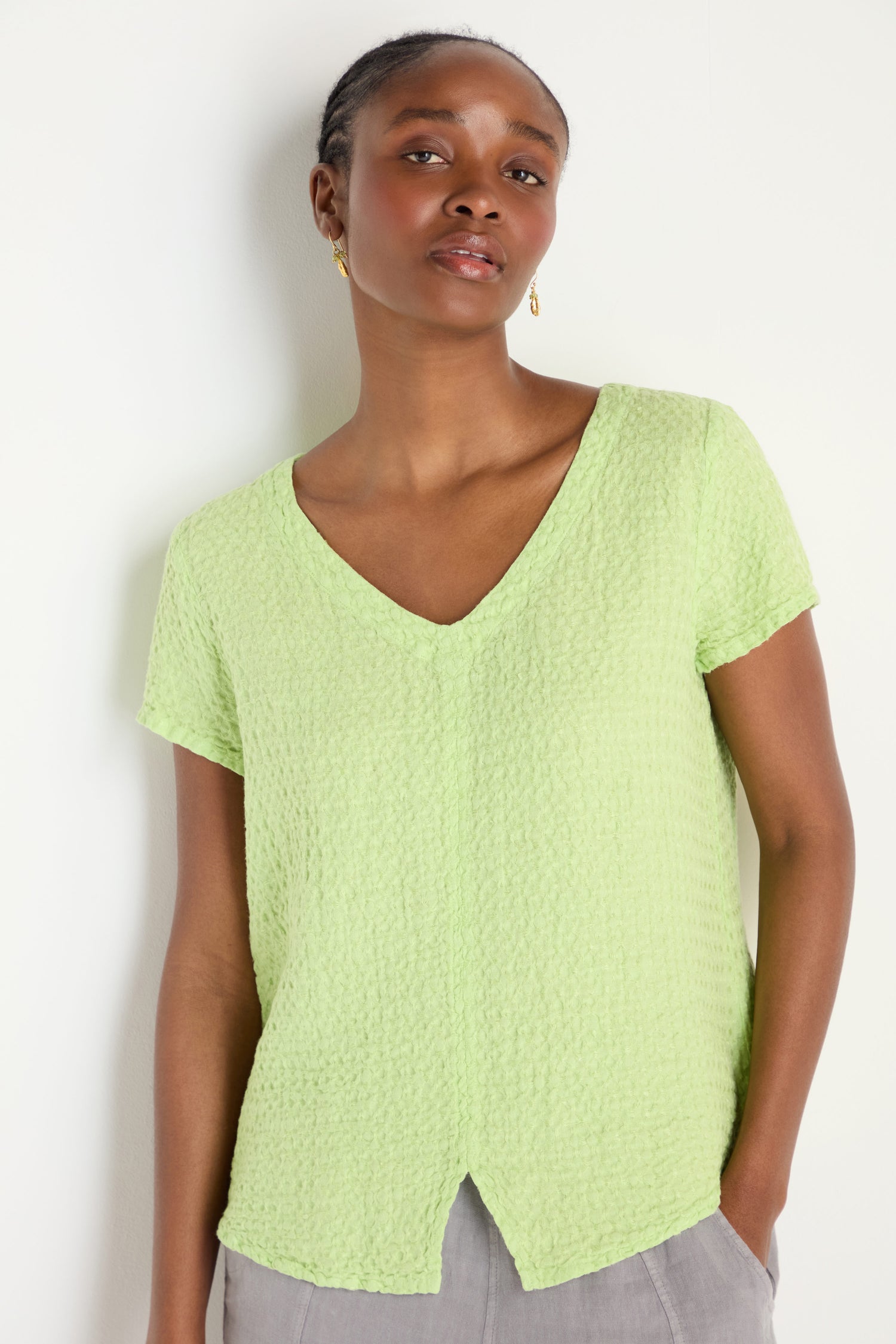 A woman stands against a white wall, wearing the Waffle Linen Lime V Neck Top with short sleeves, paired with gray pants.