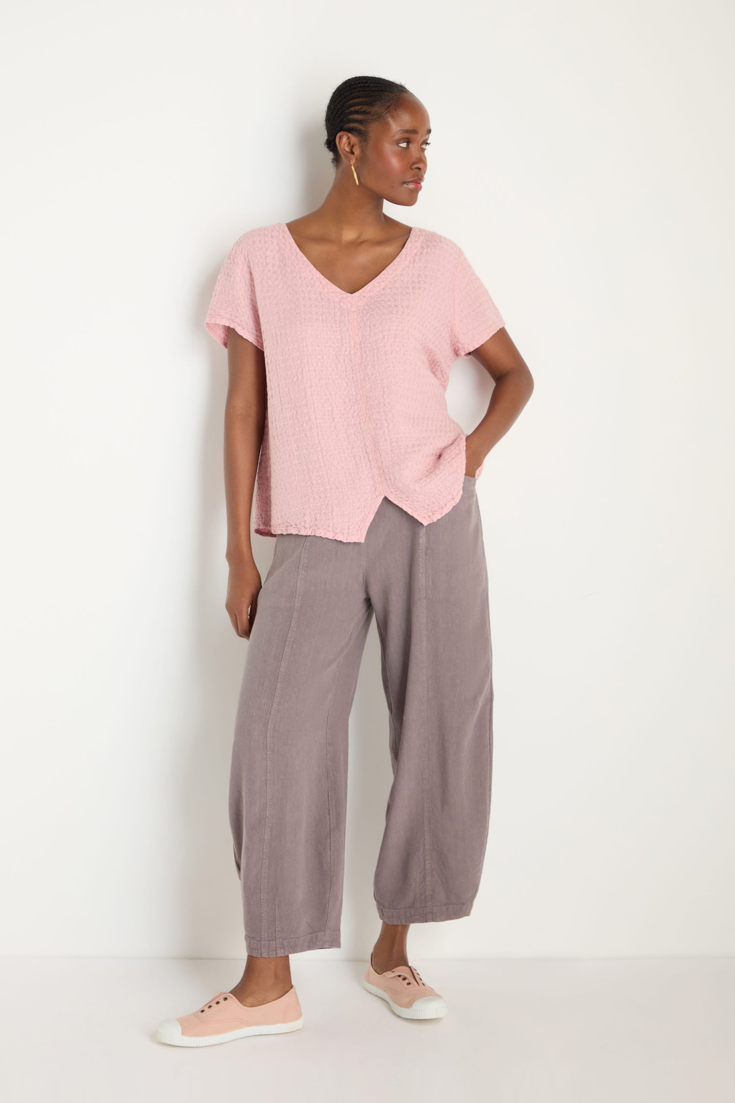 A woman stands against a white background wearing a light pink Waffle Linen V Neck Top, loose gray pants, and light pink sneakers.