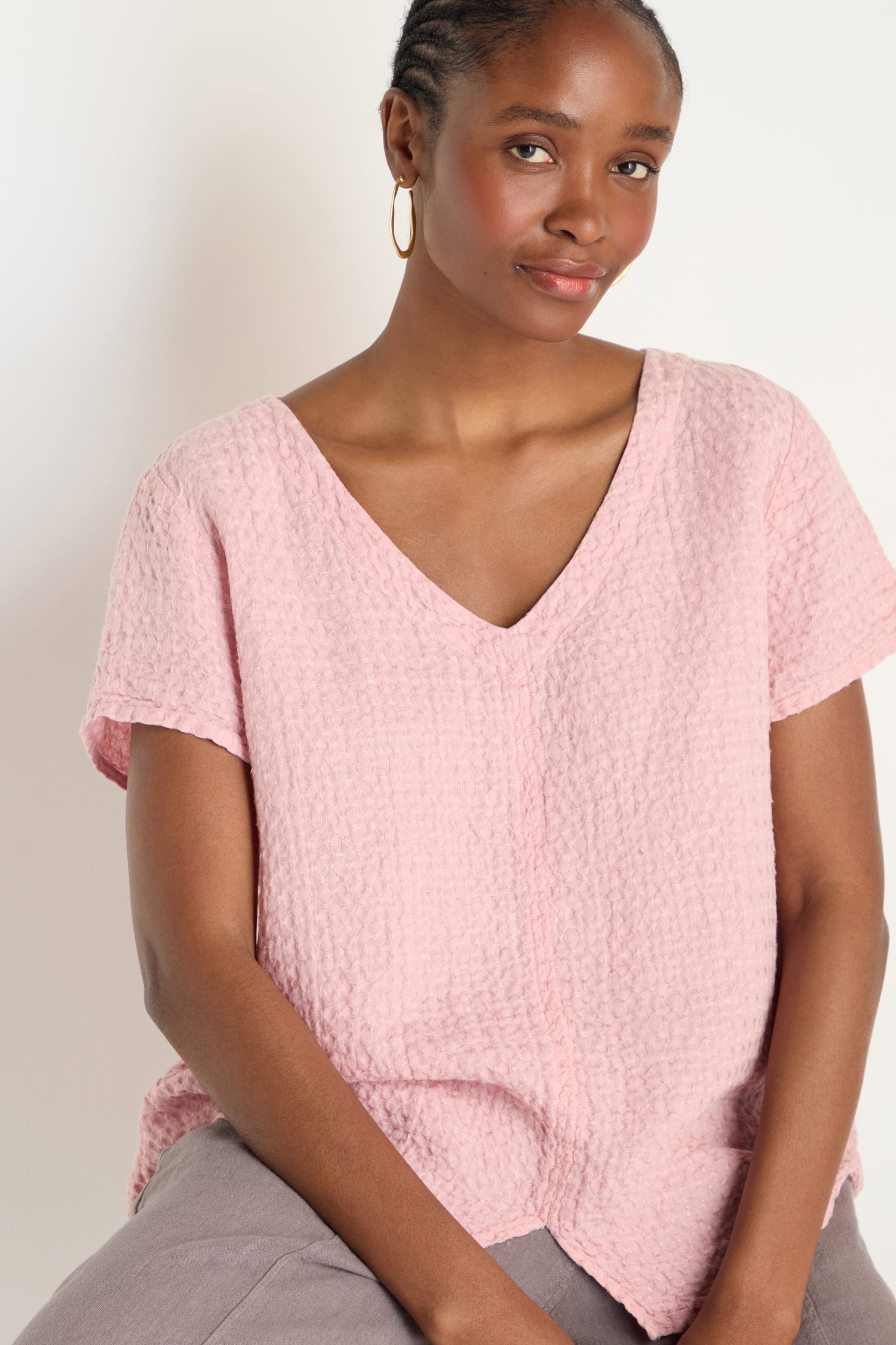 A woman sits against a light background, wearing the Waffle Linen V Neck Top in pink and gold hoop earrings from the NBEL LAT6702 collection.