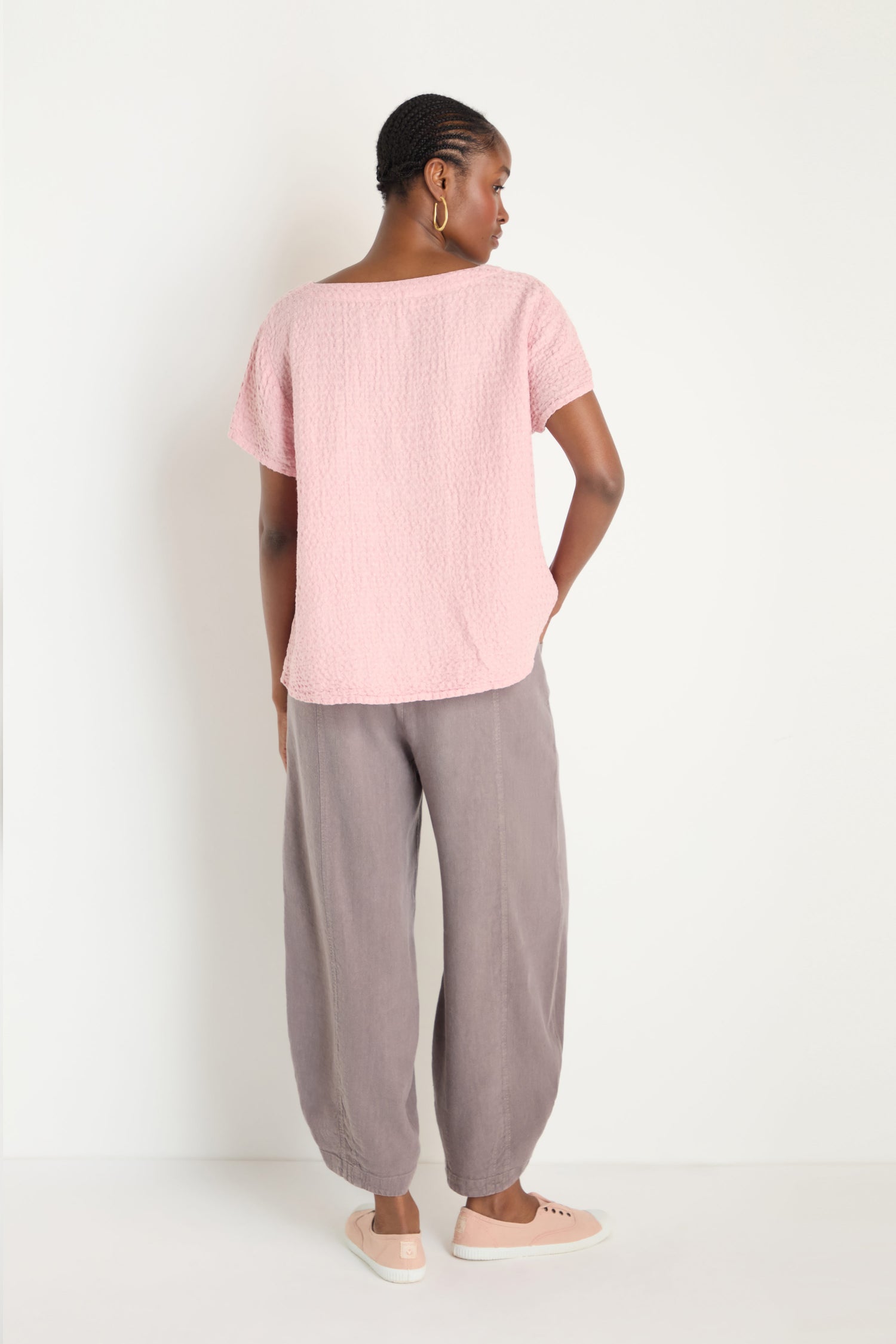 A woman faces away, wearing a textured pink Waffle Linen V Neck Top, loose grey pants, and light pink slip-on shoes against a plain white background.