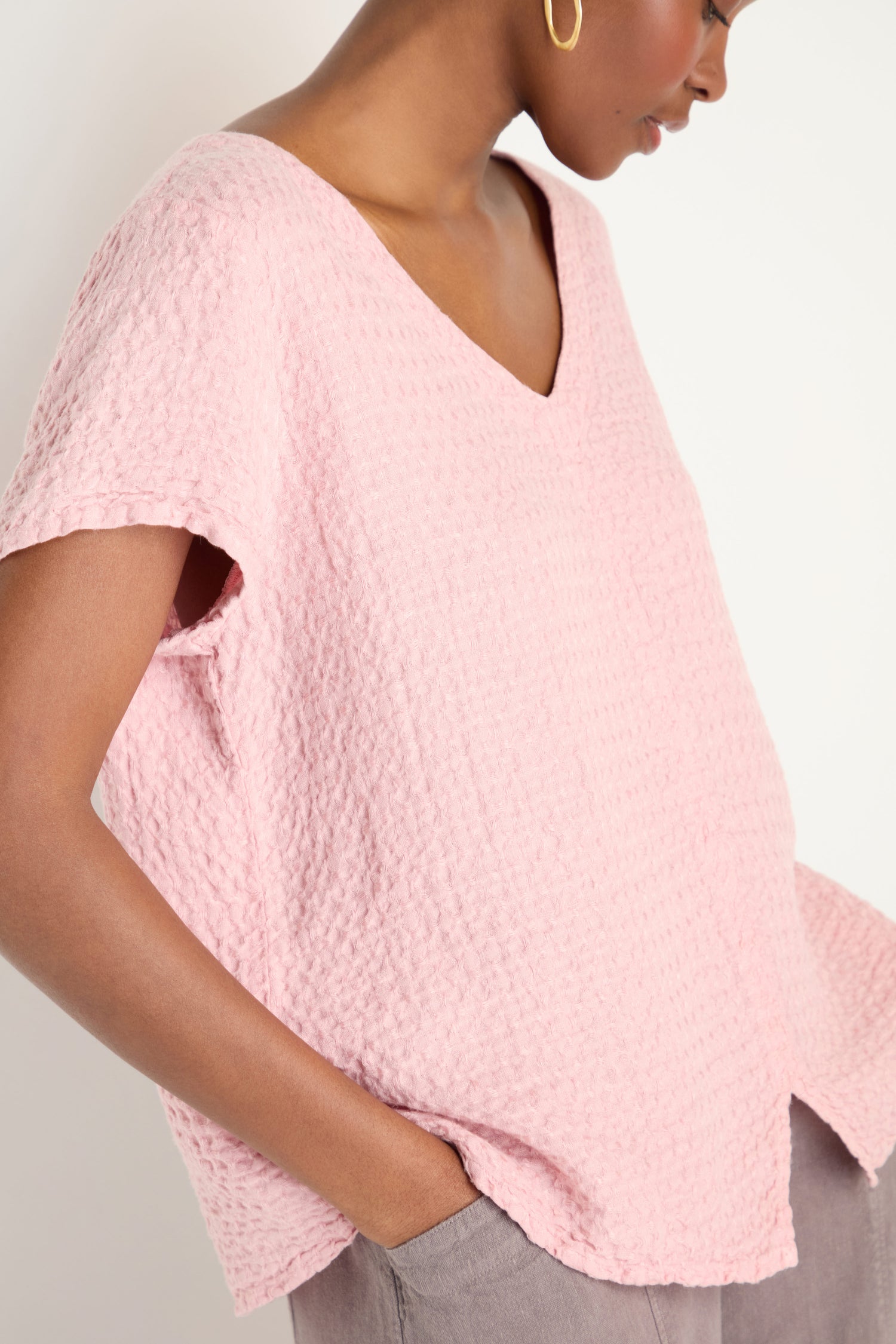 A person is wearing a textured, light pink Waffle Linen V Neck Top with short sleeves and gray pants, one hand in their pocket.