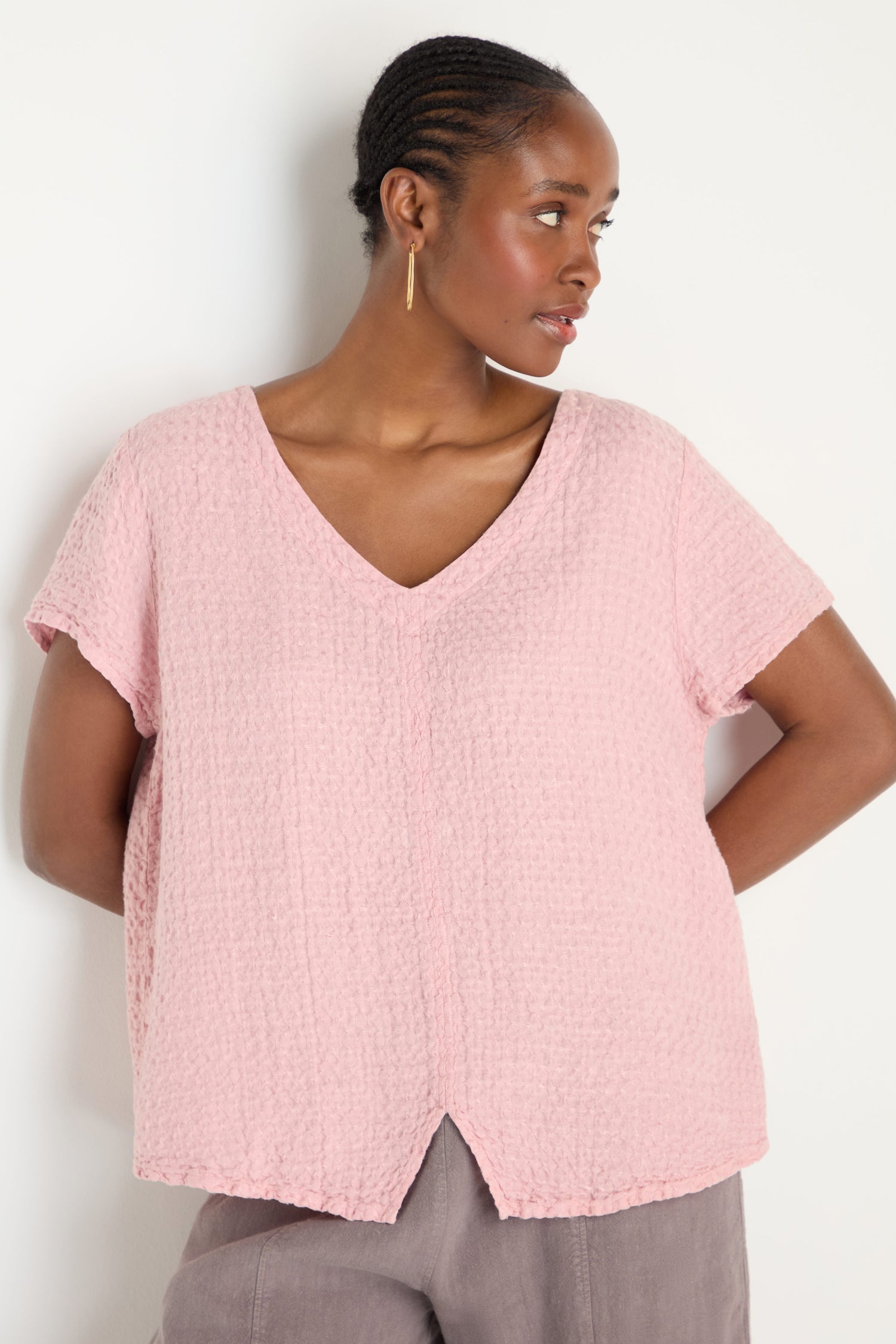 A woman in a light pink Waffle Linen V Neck Top and taupe pants stands against a white background, looking to her left.