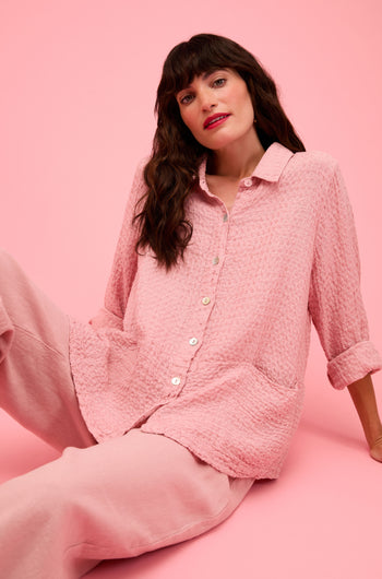 A woman with long dark hair sits against a pink background, wearing the Waffle Linen Pocket Shirt and loose pink pants—a stylish choice for relaxed layering.
