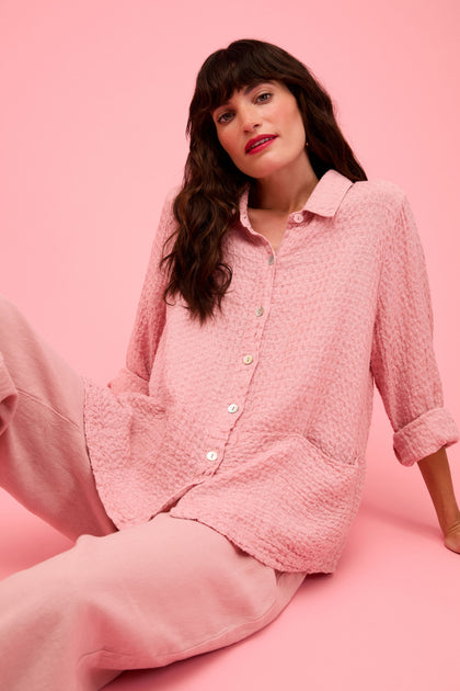 A woman with long dark hair sits against a pink background, wearing the Waffle Linen Pocket Shirt and loose pink pants—a stylish choice for relaxed layering.