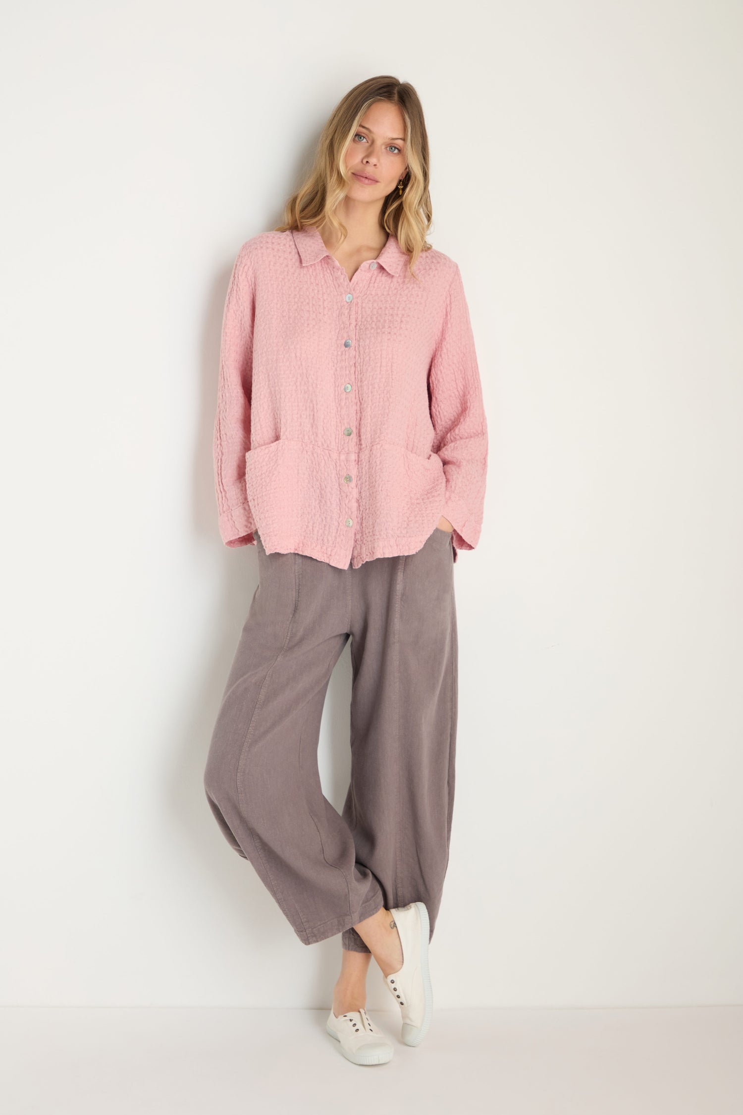 A woman stands against a white wall wearing the Waffle Linen Pocket Shirt in light pink, styled with relaxed layers, loose gray pants, and white sneakers.