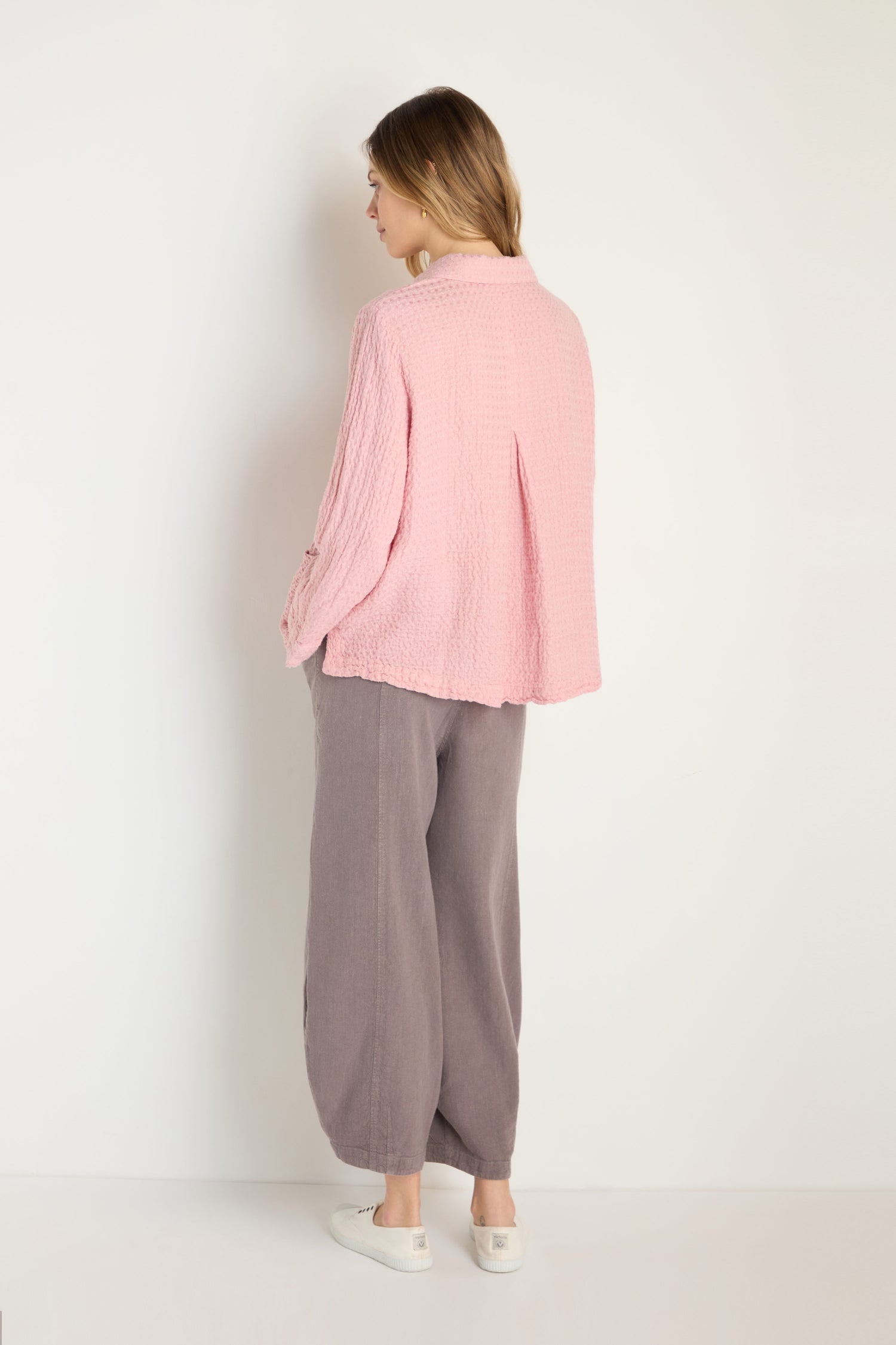A woman faces away, wearing the Waffle Linen Pocket Shirt in textured pink, loose gray pants, and white slip-on shoes—ideal for relaxed layering—set against a plain white background.