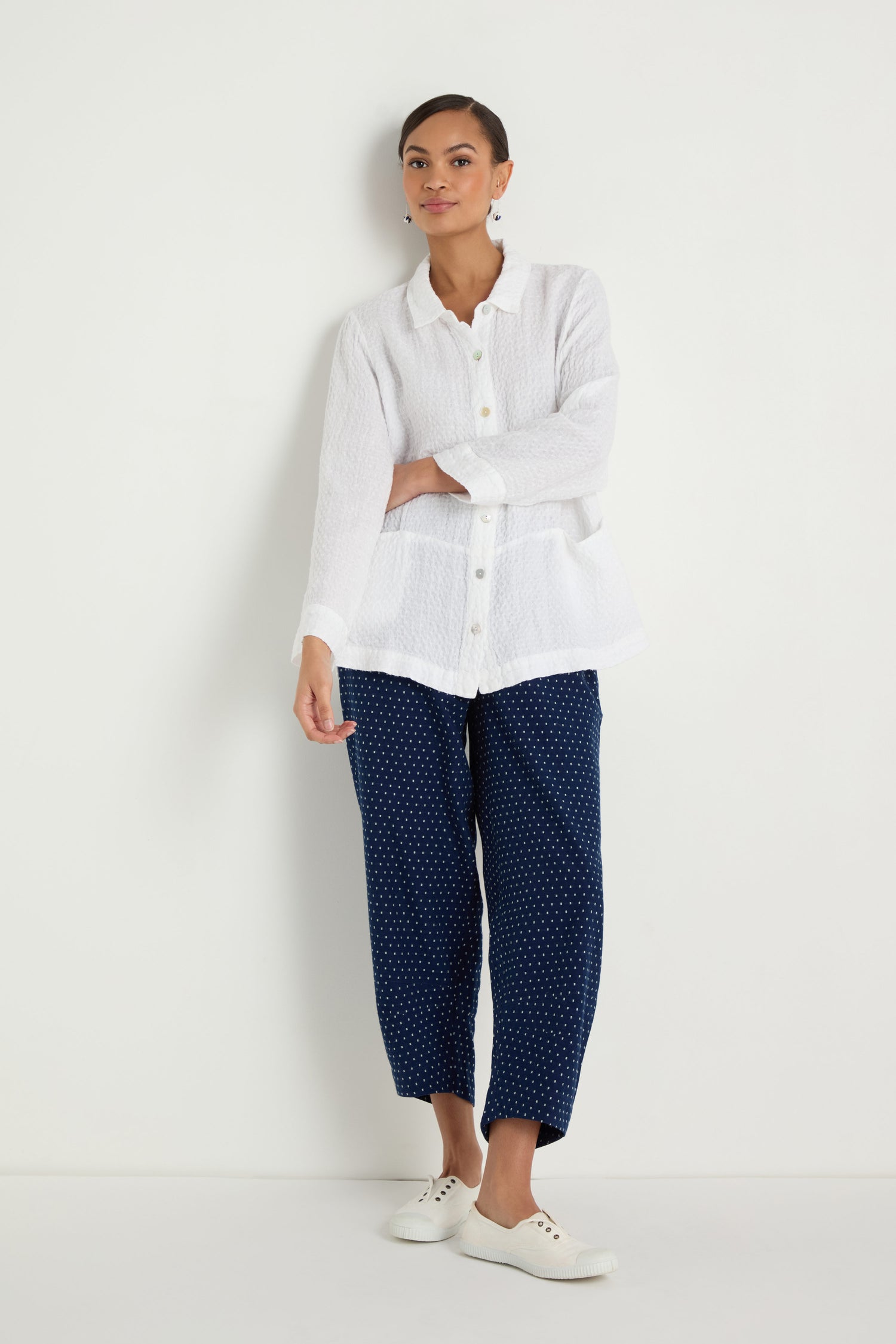 A person stands against a white wall wearing the Waffle Linen Pocket Shirt, navy blue polka dot pants, and white sneakers.