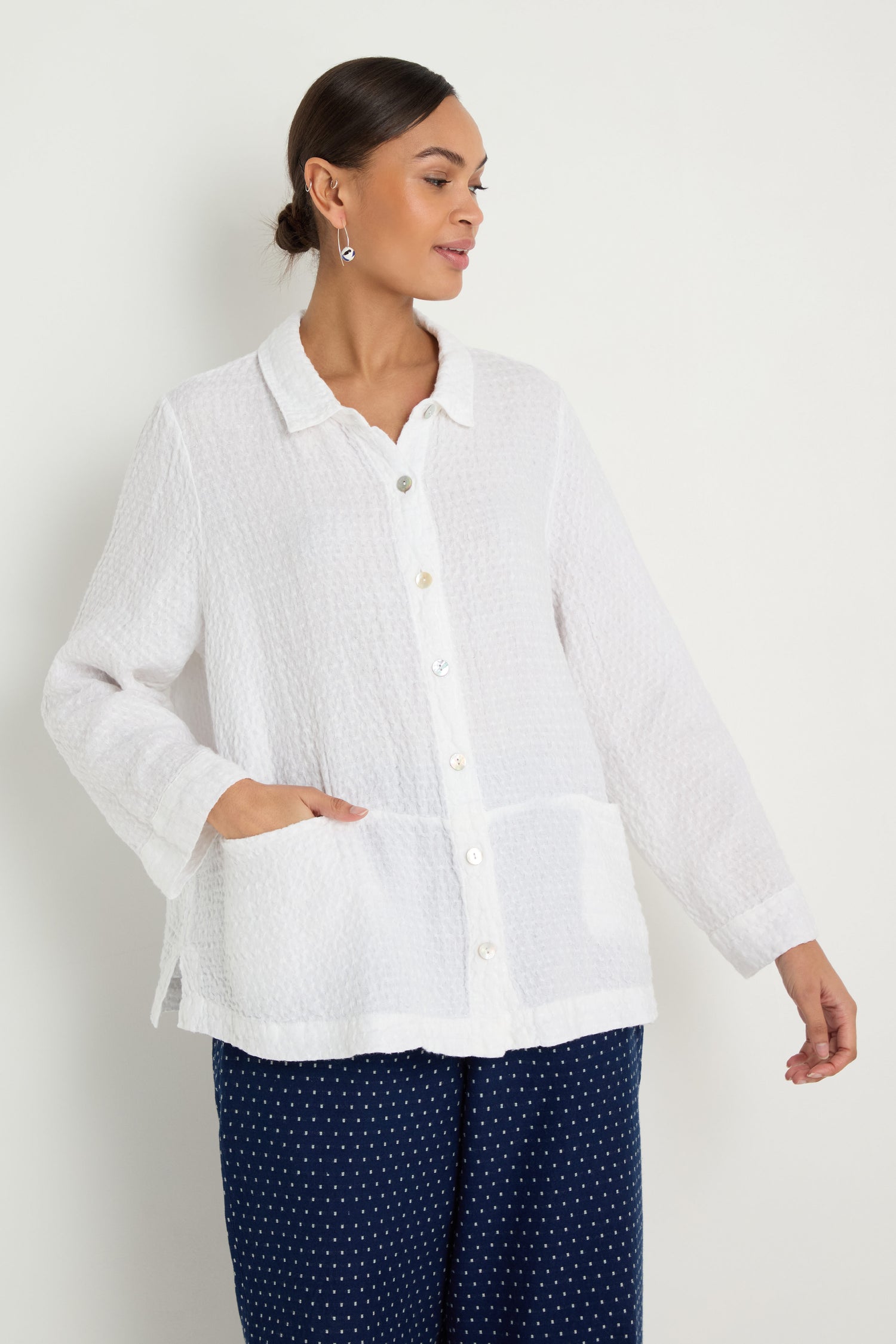A woman wears the Waffle Linen Pocket Shirt in white with a boxy silhouette and long sleeves, paired with navy blue polka dot pants, standing against a plain light background.