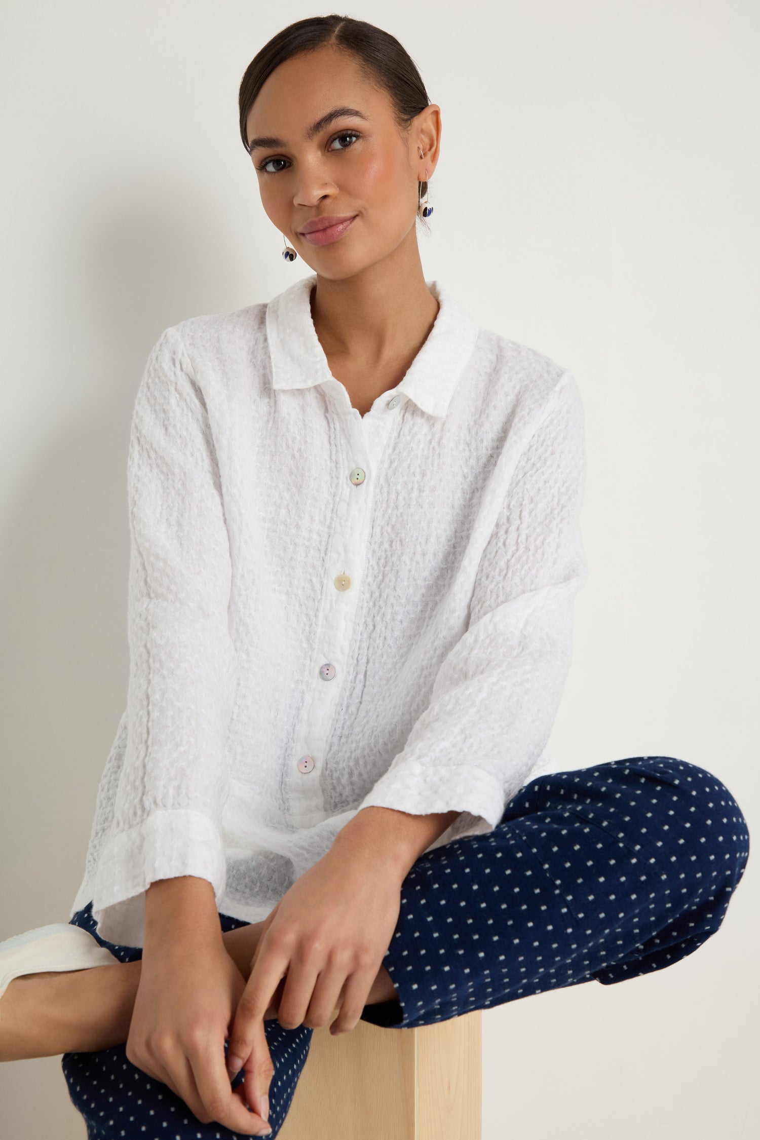A woman sits on a wooden block wearing the Waffle Linen Pocket Shirt with a boxy silhouette and navy blue dotted pants, posing against a plain light background.