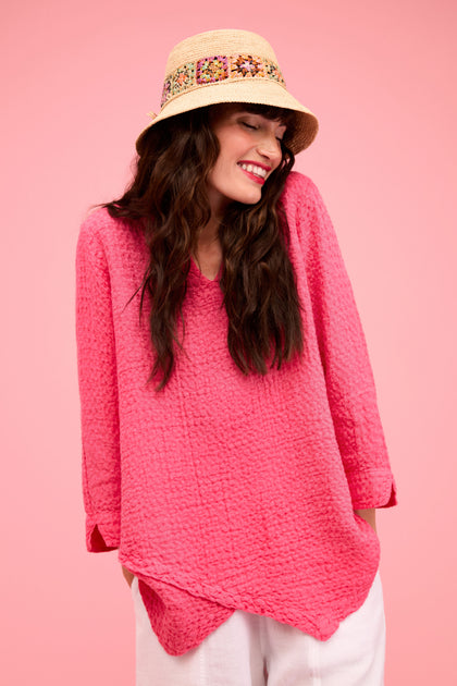 A woman smiles against a solid pink background, wearing the Waffle Linen Asymmetric Candy Tunic in pink, paired with white pants and a straw hat with a decorative band.