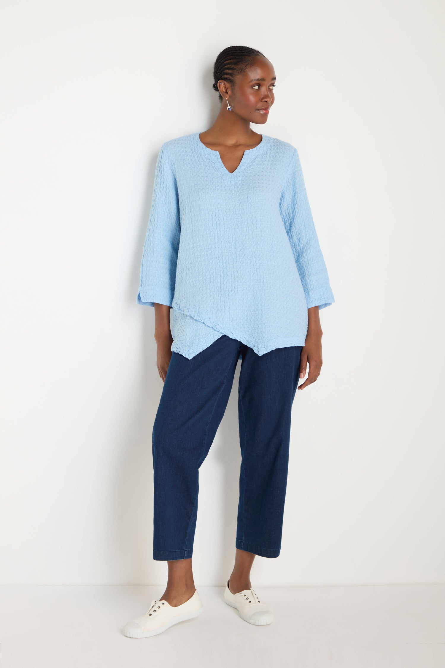 A woman stands against a white wall wearing the Waffle Linen Asymmetric Tunic in light blue, paired with dark blue cropped pants and white sneakers.