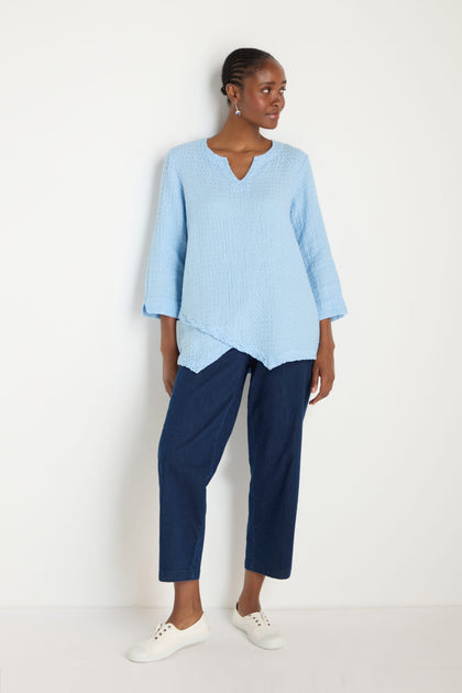 A woman stands against a white wall wearing the Waffle Linen Asymmetric Tunic in light blue, paired with dark blue cropped pants and white sneakers.