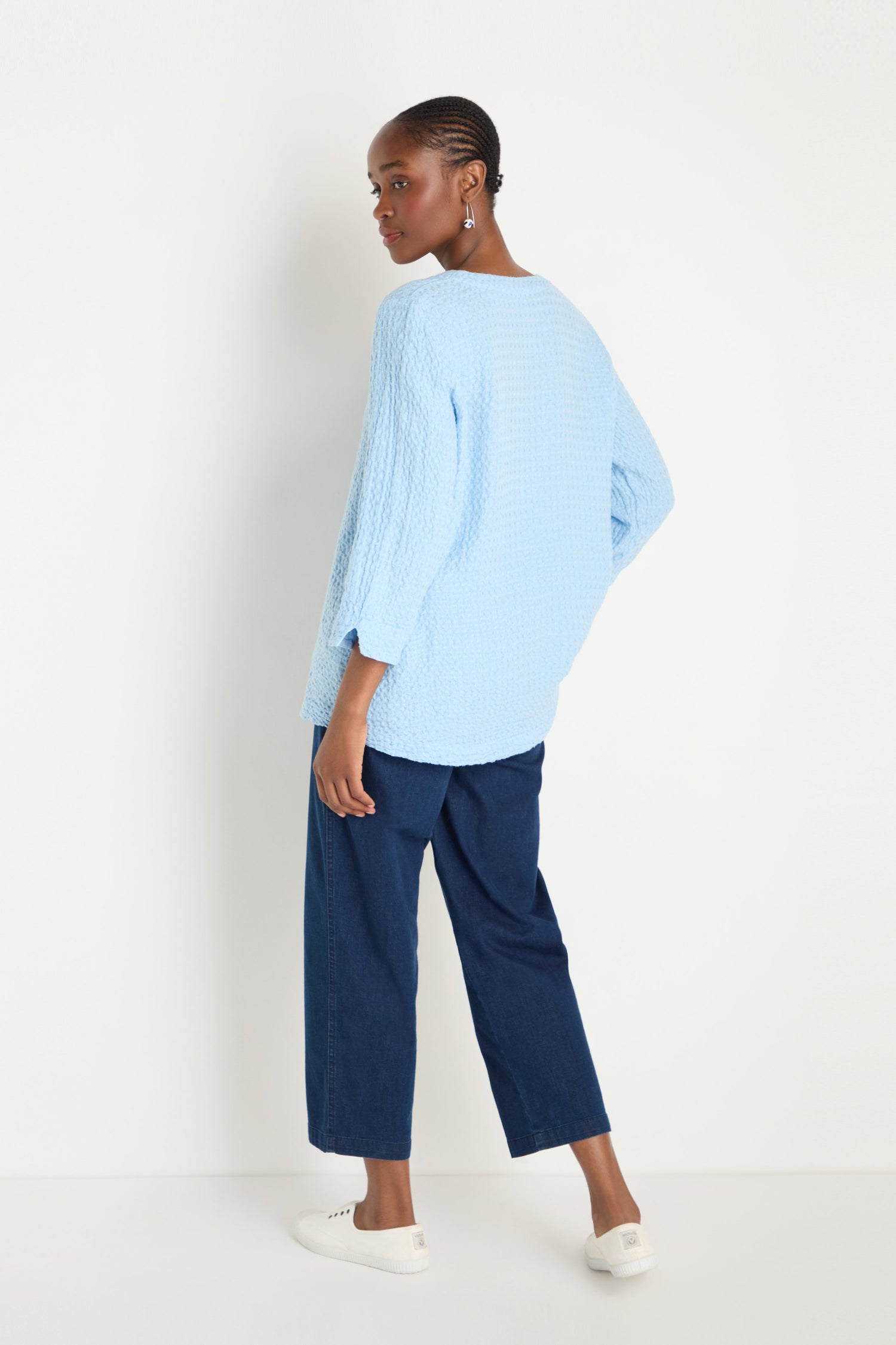 A woman stands against a white wall wearing the Waffle Linen Asymmetric Tunic in light blue, paired with dark blue wide-leg jeans and white slip-on shoes, looking over her shoulder.