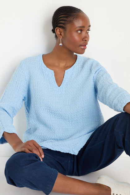 A woman in a light blue Waffle Linen Asymmetric Tunic and dark blue pants sits on the floor against a white background, looking to her left.
