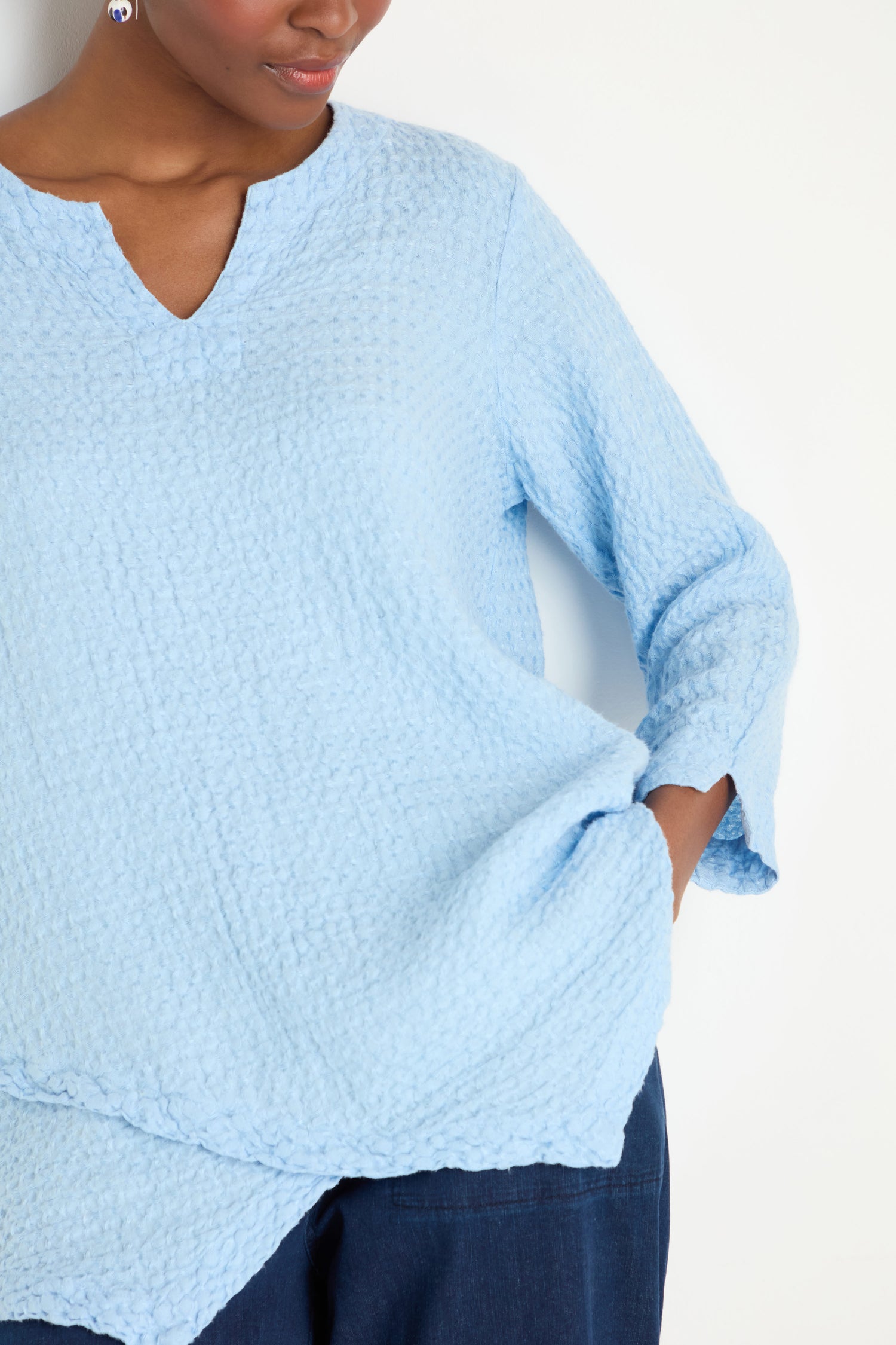 A person wears the Waffle Linen Asymmetric Tunic in light blue with long sleeves and dark pants, standing against a white background with one hand in their pocket.