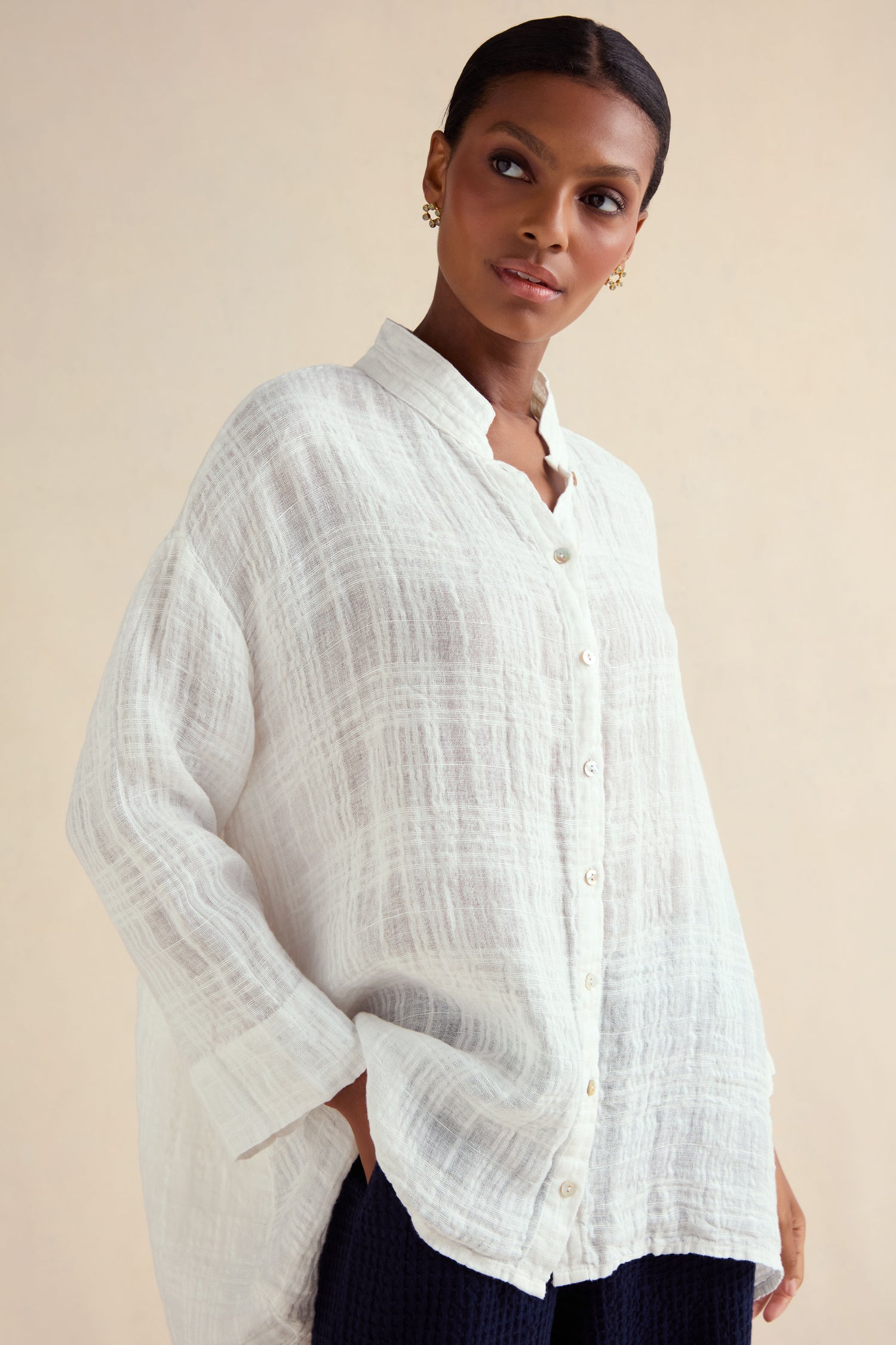 Woman wearing the Gauzy Linen Relaxed Shirt in white with a subtle woven check texture, paired with dark pants and gold earrings, standing against a light background.
