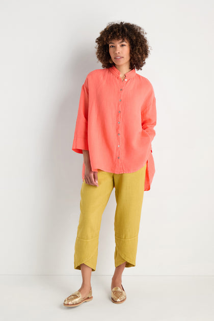 A person stands against a plain white background, wearing the Organza Linen Flared Shirt in coral, paired with yellow cropped pants and woven slip-on shoes.