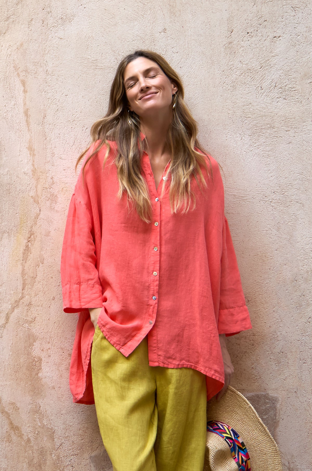 A woman stands against a textured beige wall, wearing a loose coral Organza Linen Flared Shirt and yellow pants, one hand in her pocket, the other holding a straw hat, eyes closed and smiling softly.