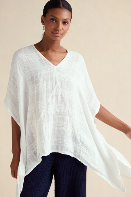 A woman stands against a plain beige background wearing the Gauzy Linen Boxy Tunic, a loose-fitting, oversized white V-neck top, paired with dark pants.