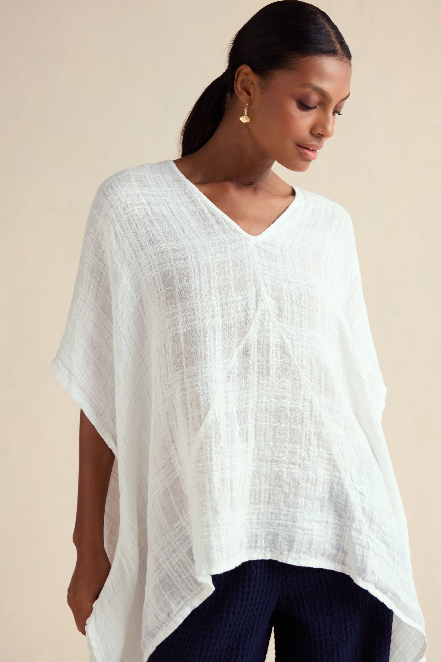 A person wearing the Gauzy Linen Boxy Tunic, a white textured V-neck top with an oversized fit, paired with dark pants, gold earrings, and hair tied back, stands against a neutral background.