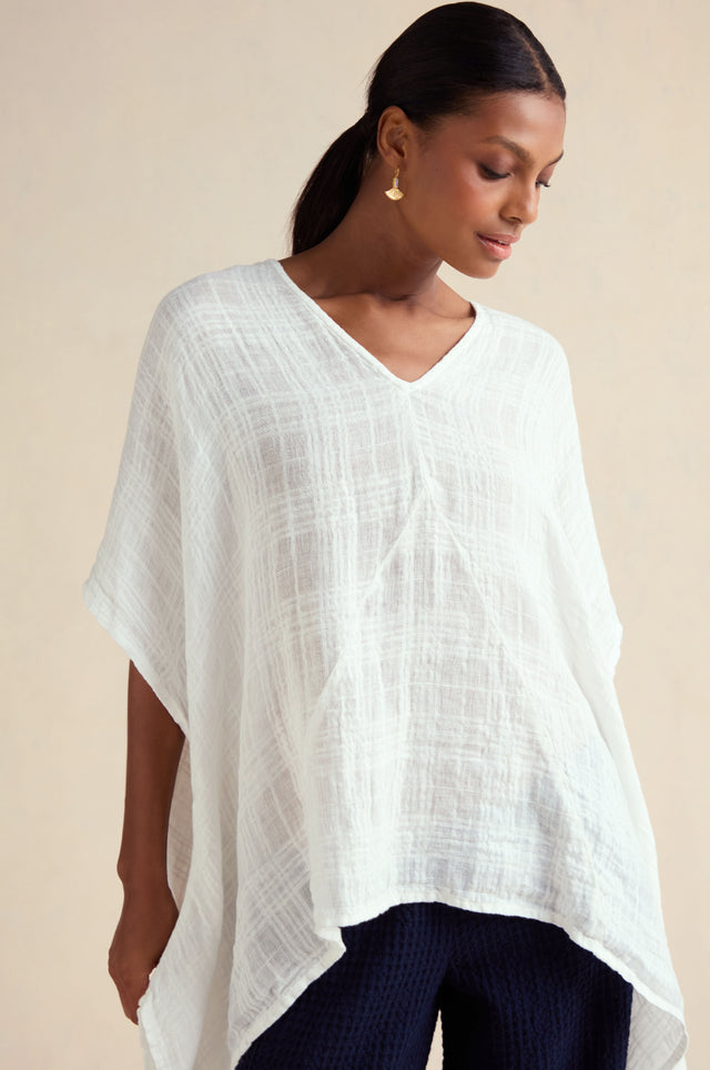 A person wearing the Gauzy Linen Boxy Tunic, a white textured V-neck top with an oversized fit, paired with dark pants, gold earrings, and hair tied back, stands against a neutral background.