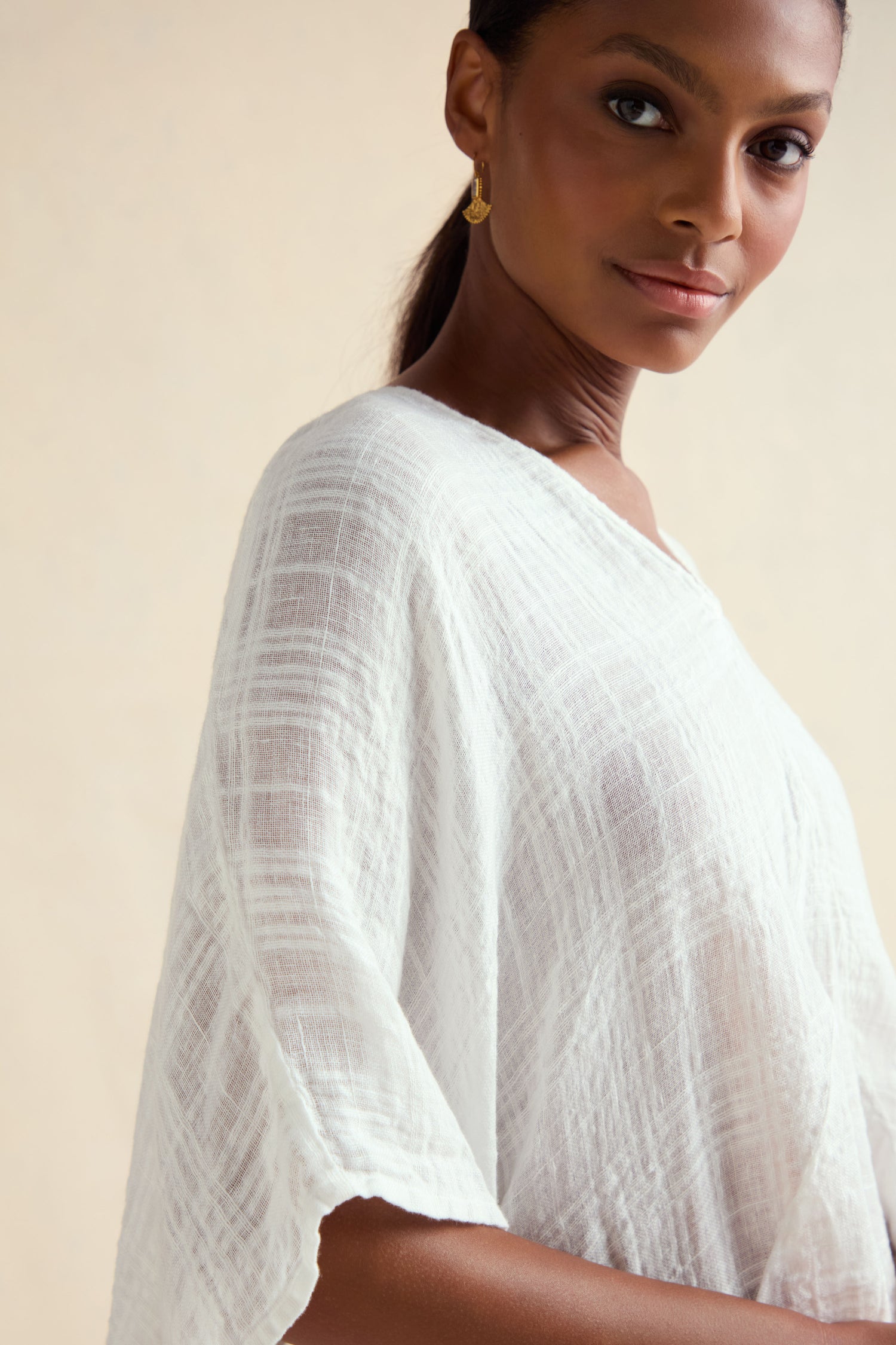 A woman stands against a light beige background, looking slightly toward the camera, wearing the Gauzy Linen Boxy Tunic in white with a textured finish and oversized fit, paired with gold hoop earrings.