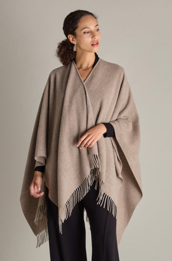 Draped in the Wool Cashmere Poncho with fringe detailing, an individual stands before a neutral backdrop.