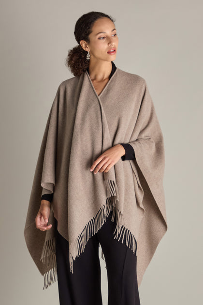 Draped in the Wool Cashmere Poncho with fringe detailing, an individual stands before a neutral backdrop.