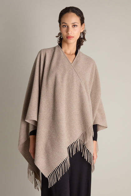 A person is adorned in the Wool Cashmere Poncho, featuring elegant fringe detailing, layered over a black outfit and posed against a neutral background.