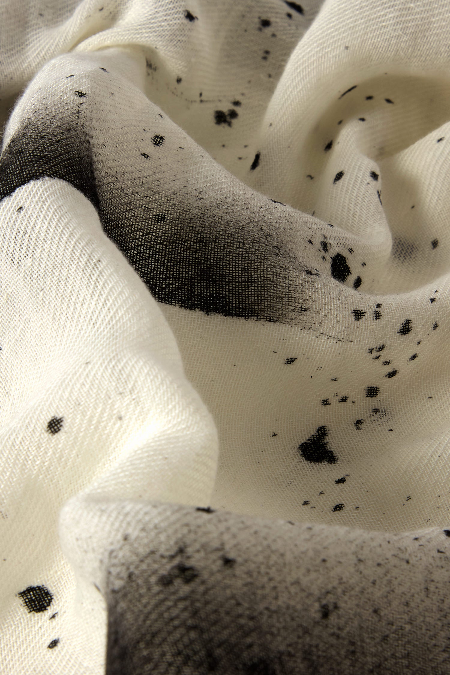 Close-up of white fabric with scattered black ink blots and a textured surface, resembling the artistic flair of the Abstract Brushstroke Scarf, perfect for adding creative charm to your spring wardrobe.